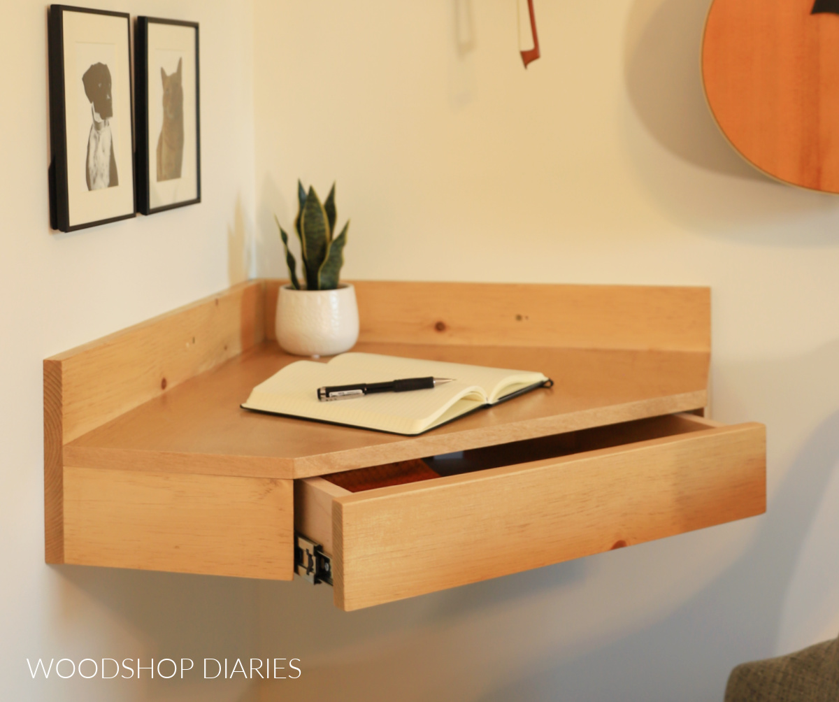 Floating wooden corner desk with drawer open hanging on wall with open notebook