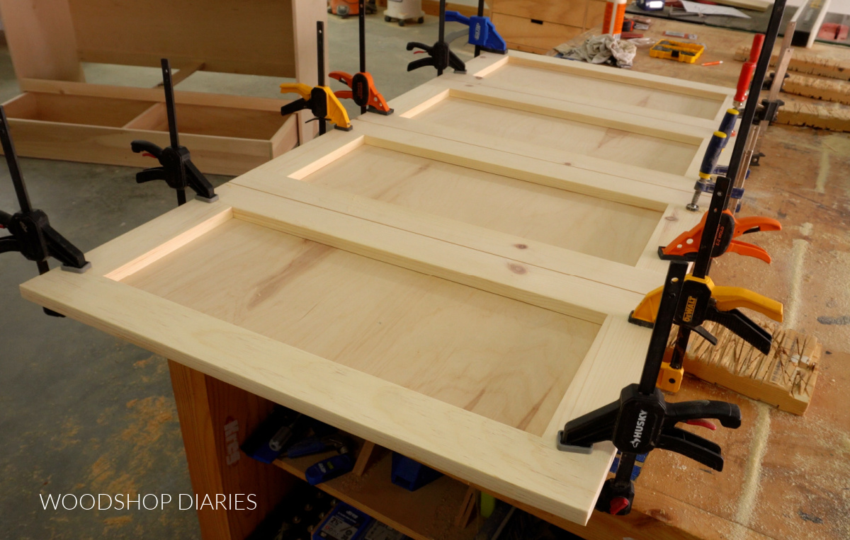 Faux door frames glued and clamped onto plywood panel on workbench