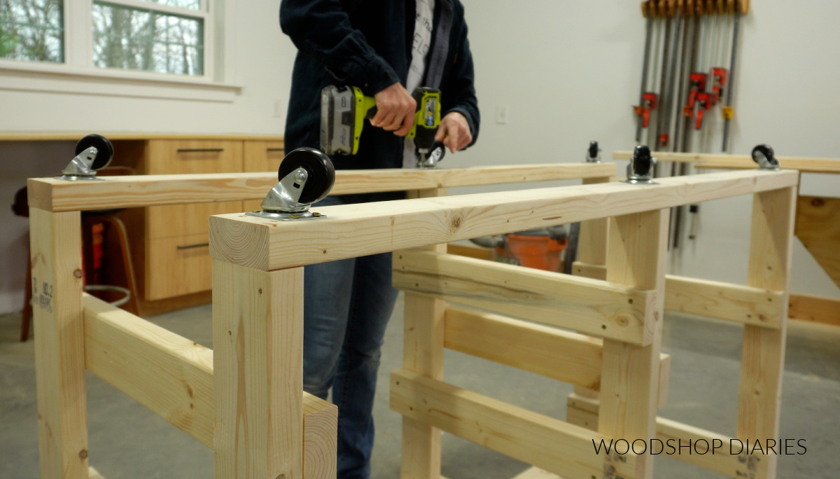 Shara Woodshop Diaries screwing caster wheels onto bottom of workbench frame