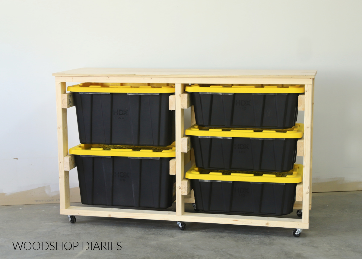 Completed DIY tote storage workbench frame made with 2x4s and a plywood top--five black totes with yellow lids inside frame