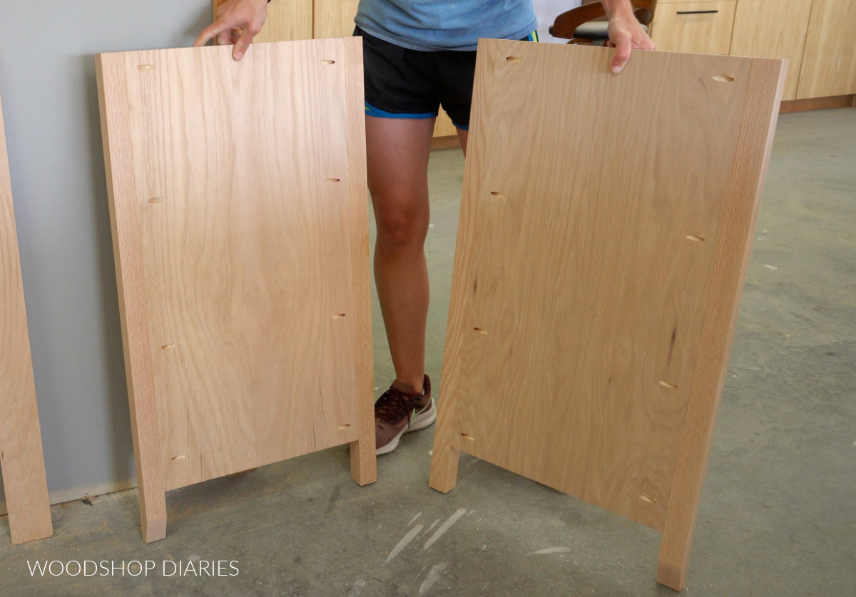Shara Woodshop Diaries holding two assembled side panels for DIY single sink bathroom vanity made with red oak