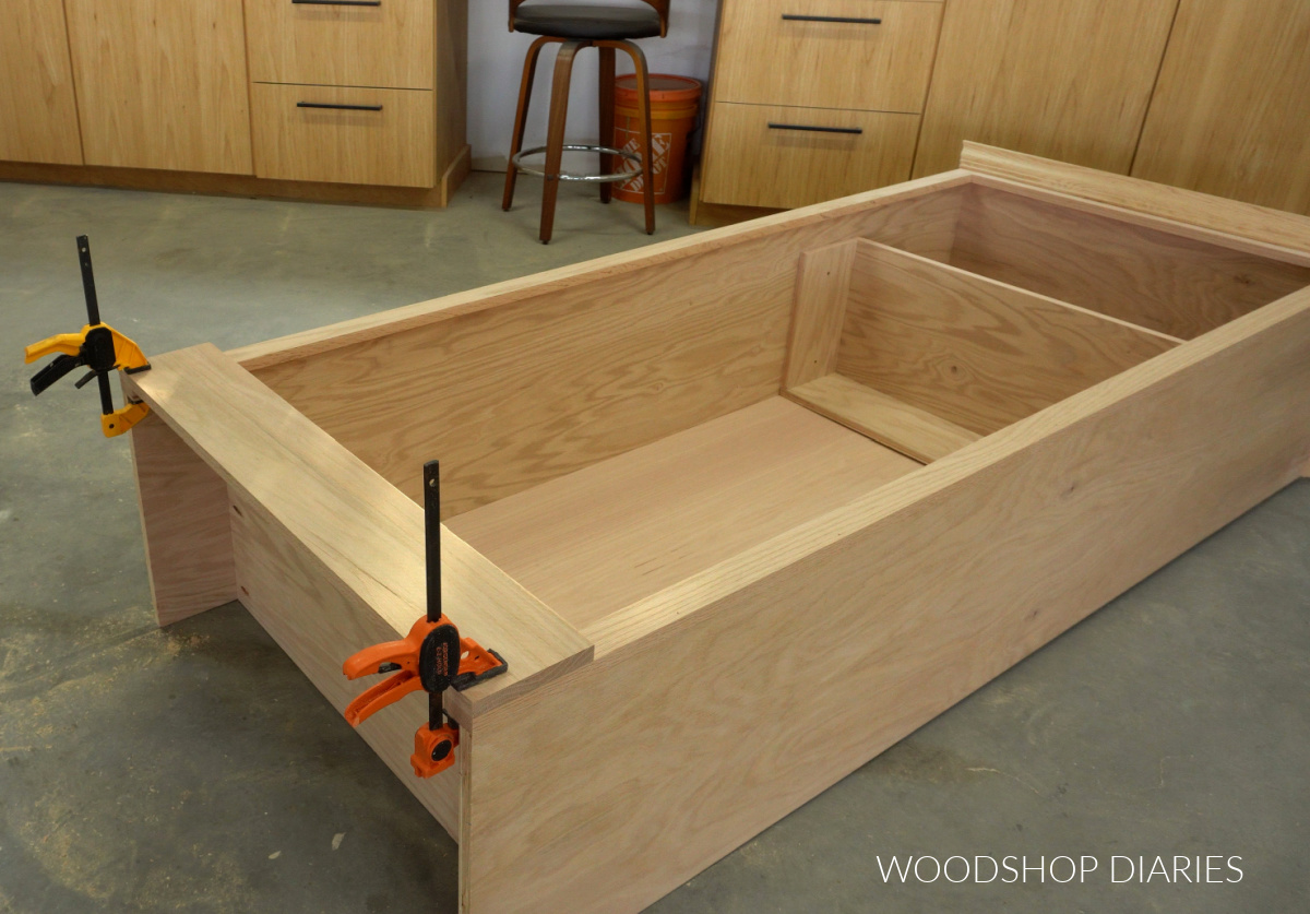 Tall coat storage cabinet laid on its back on workshop floor with crown molding and 1x6 baseboard trim clamped onto bottom