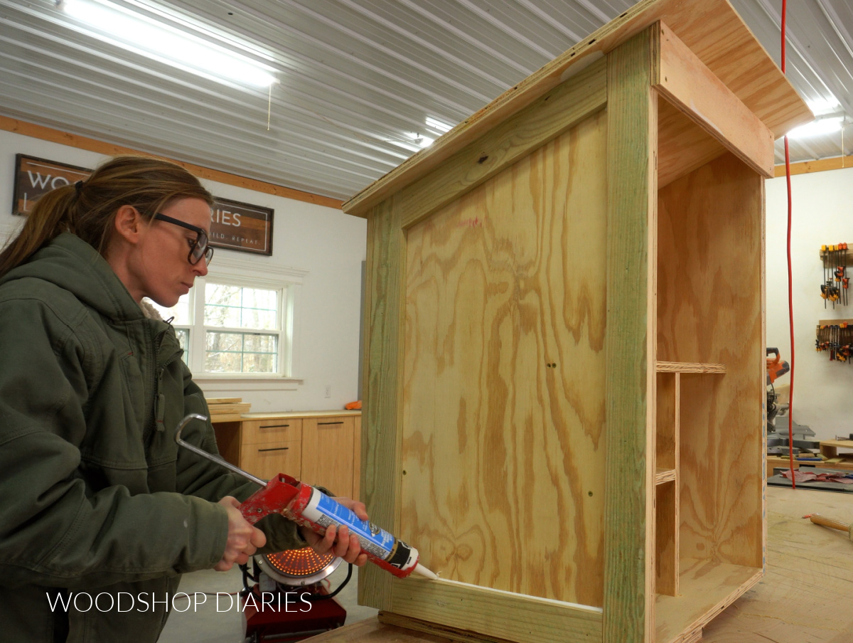 Shara Woodshop Diaries applying caulk to trim on side of free little pantry box on workshop