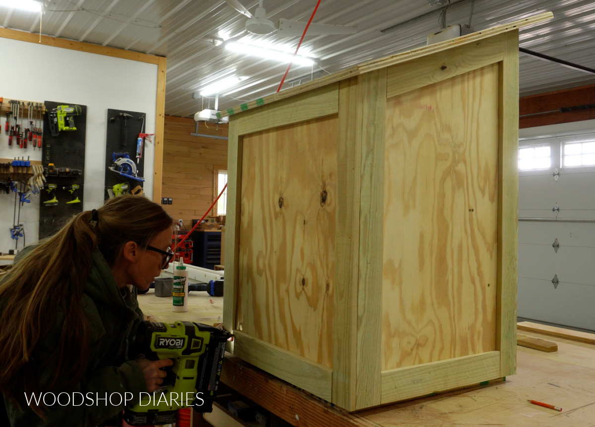 Shara Woodshop Diaries using brad nailer to secure trim pieces to back of community blessing box on workbench