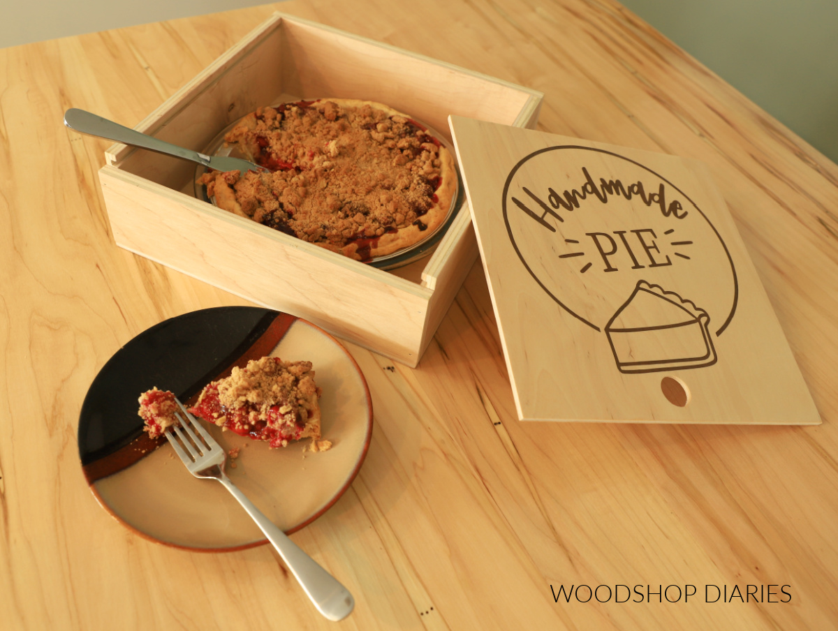 Wooden pie box with sliding lid removed. Cherry pie inside with a slice cut on a plate