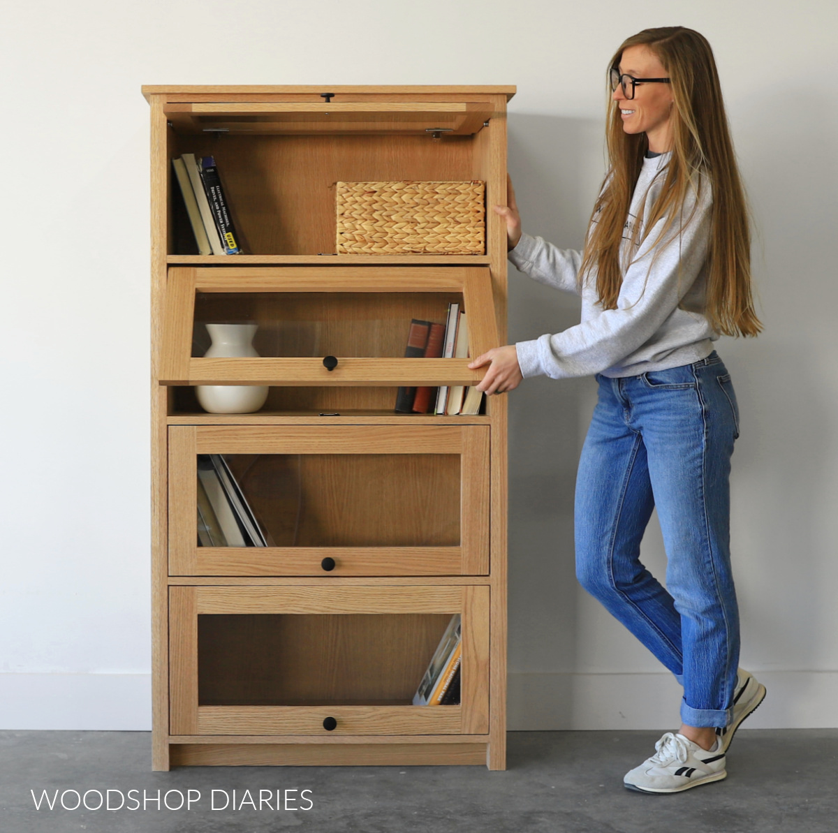 Shara Woodshop Diaries standing next to DIY lawyer bookcase opening second door from the top. Top door open and bottom two doors closed. Bookcase made from red oak