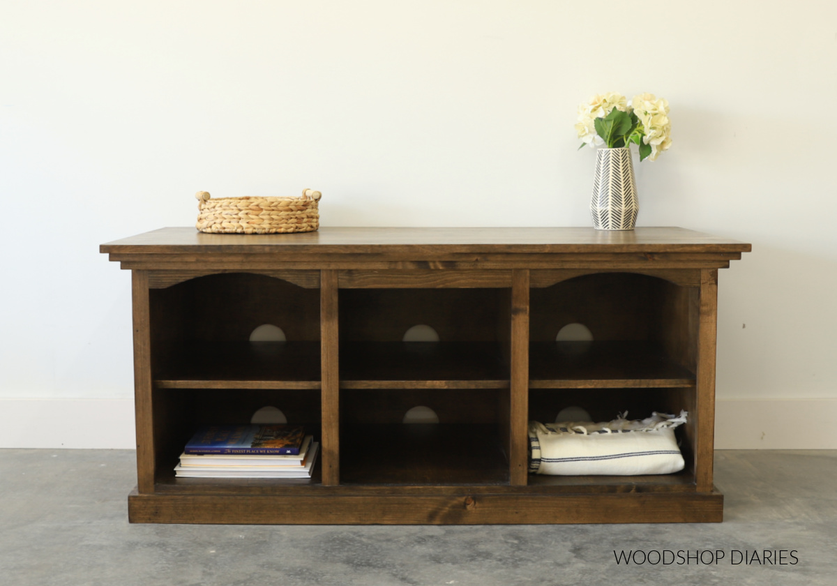 Completed DIY TV stand with six open cubbies and holes in the back for cords and cables. Stained dark brown with books and blankets inside
