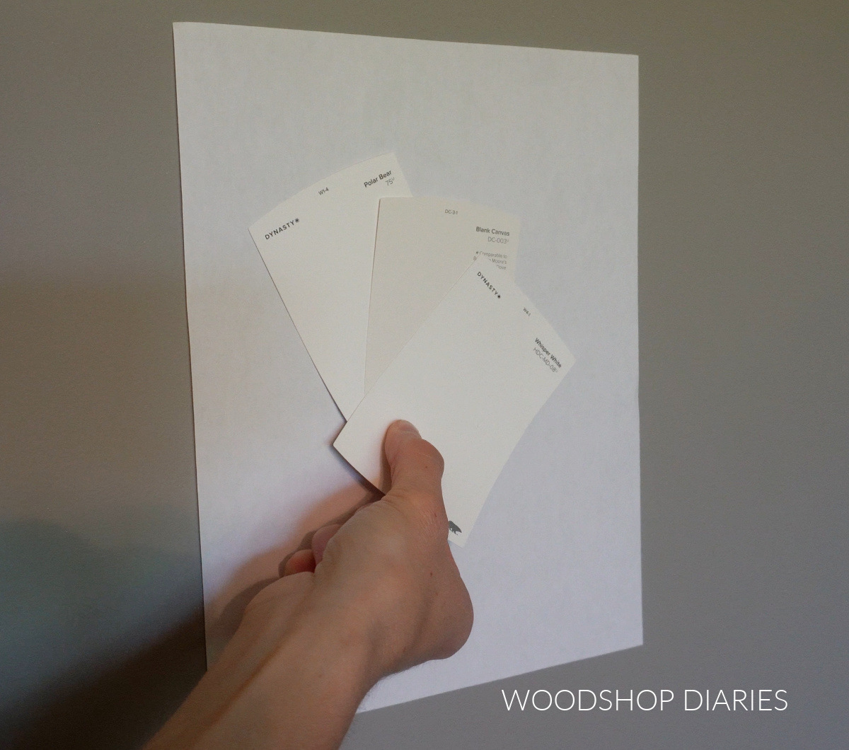 Shara Woodshop Diaries holding up three BEHR white paint color shades against white paper on bedroom wall to show undertones