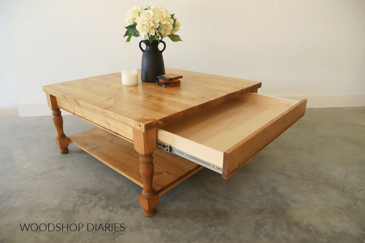 large square wooden coffee table with bottom shelf, turned legs and drawer. Drawer is open 